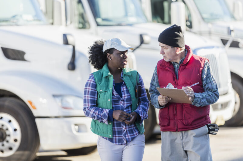 Two multi-ethnic truck drivers, a senior Hispanic man in his 60s and a mid-adult African-American woman in her 30s, standing in front of a fleet of semi-trucks or tractor-trailers. The man is holding a clipboard and they are conversing, looking at each other face to face.