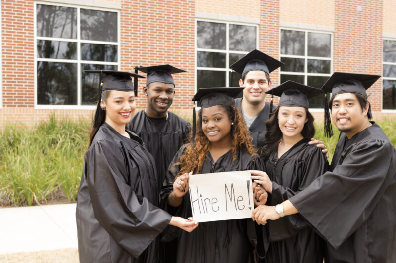 Six multi-ethnic graduates hold 'Hire Me' sign after college graduation. School building background. Employment issues concept.