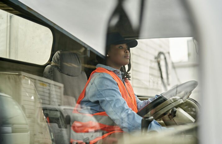 Personal perspective of mid 20s woman in casual clothing, cap, and reflective vest sitting in driver’s seat, looking straight ahead, ready to begin road trip.