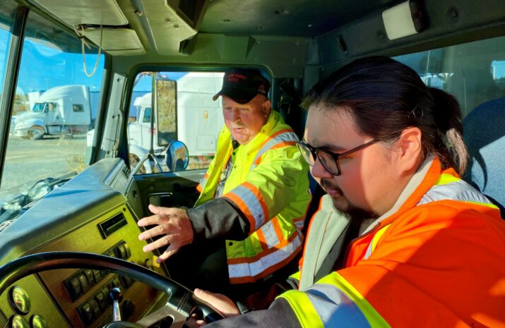 A trainer instructs a student in a truck cab.