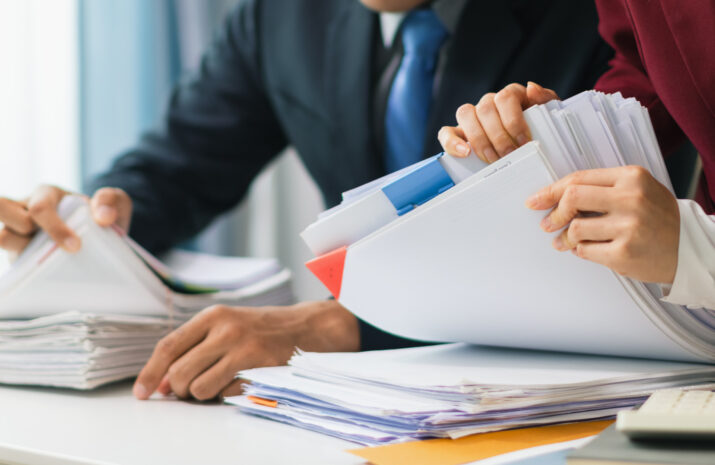 Two businesswomen are working together to search through important documents from a pile of documents on a table to verify the authenticity of the documents that were previously signed in a contract.