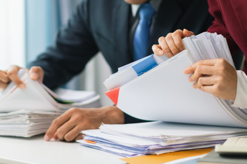 Two businesswomen are working together to search through important documents from a pile of documents on a table to verify the authenticity of the documents that were previously signed in a contract.